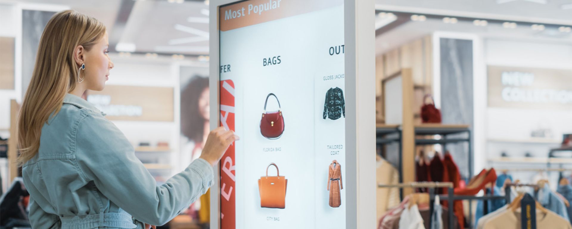 A woman in a denim jacket examines a digital display featuring popular bags in a modern store, enhancing the tangible user experience, with blurred shoppers in the background.