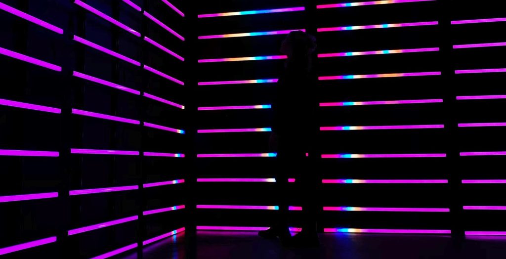 Silhouetted figure stands among glowing pink and purple neon light bars in a dark room, creating a futuristic, phygital atmosphere.