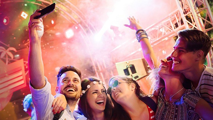 A group of four friends enjoying a concert, laughing and taking selfies with colorful stage lights and smoke in the background, experiencing the blend of physical and digital in a phygital environment.
