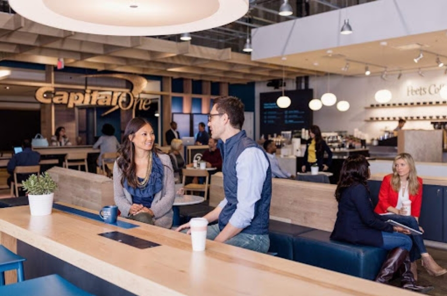 A man and a woman engaging in a conversation at a café counter inside a spacious, modern area with the "Capital One" and "Peet's Coffee" logos in the background, enhancing the int