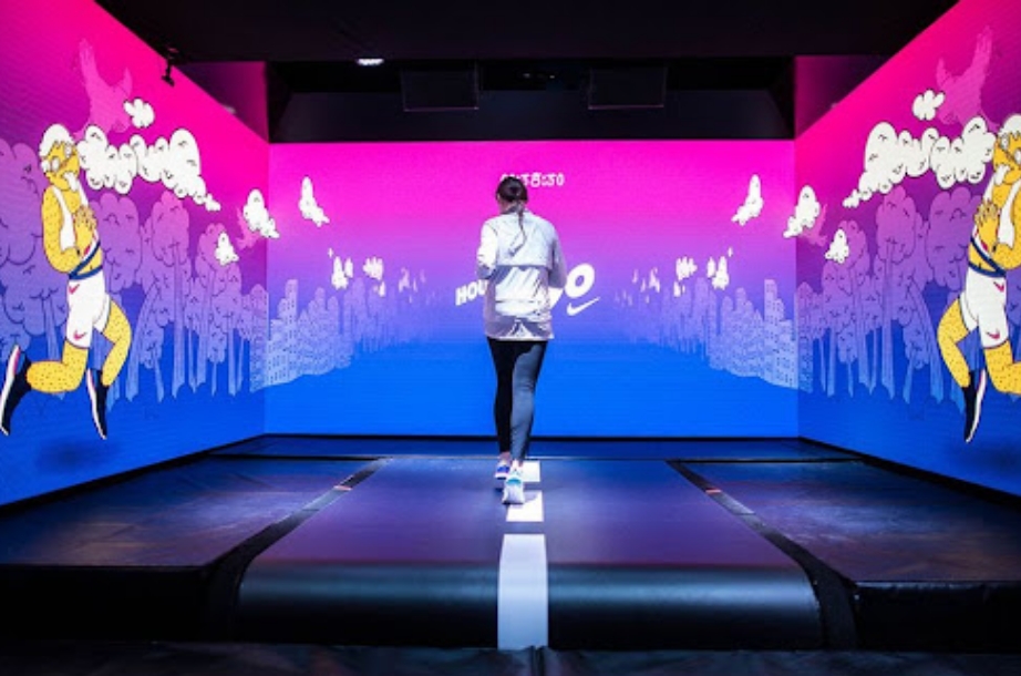 A person walks on a treadmill in a vibrant room illuminated in purple light, featuring large cartoon-style illustrations of exercising characters on the surrounding walls, enhancing the intangible user experience.