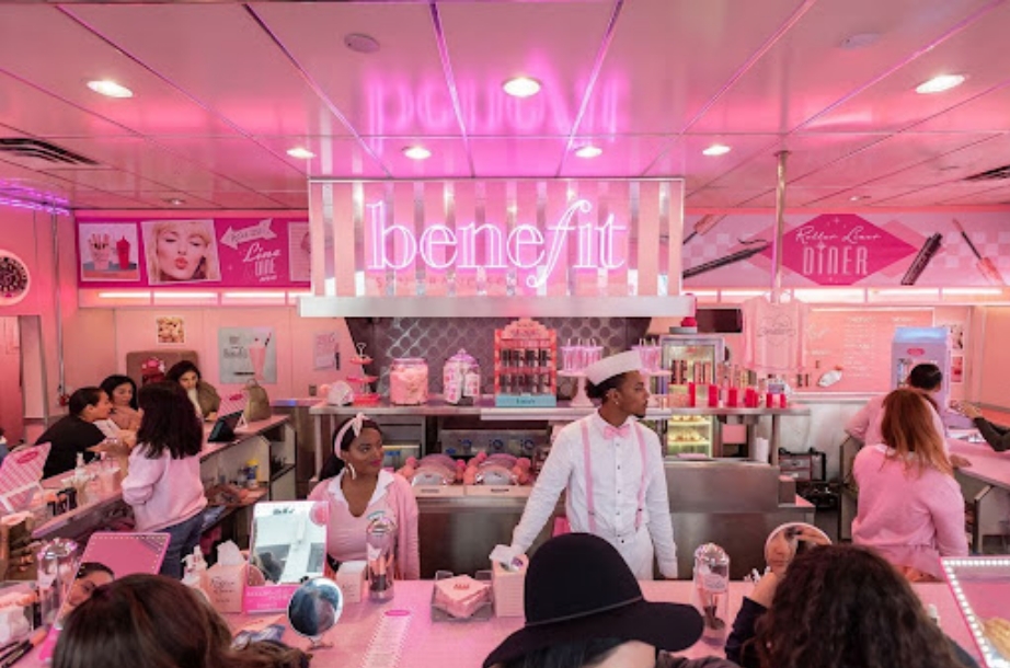 Image of a lively Benefit Cosmetics store with pink neon signs, featuring shoppers exploring beauty products and staff assisting them to enhance user experience. The interior is vibrant and stylish, decorated in shades of pink.