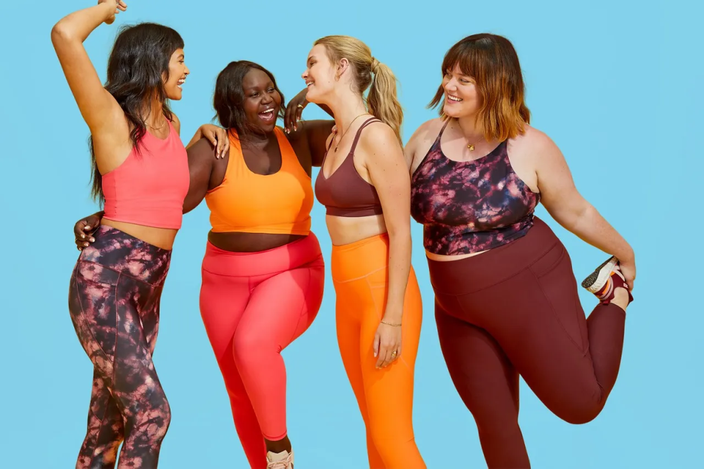 Four women of various body types, wearing colorful activewear, joyfully posing together against a light blue background. They appear happy and confident as they influentially promote the brand.