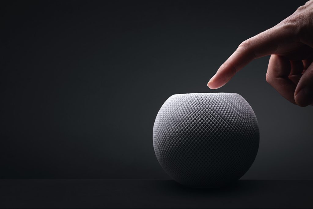 A finger about to touch a textured, spherical smart home speaker on a dark background, highlighting the interaction between human and device.