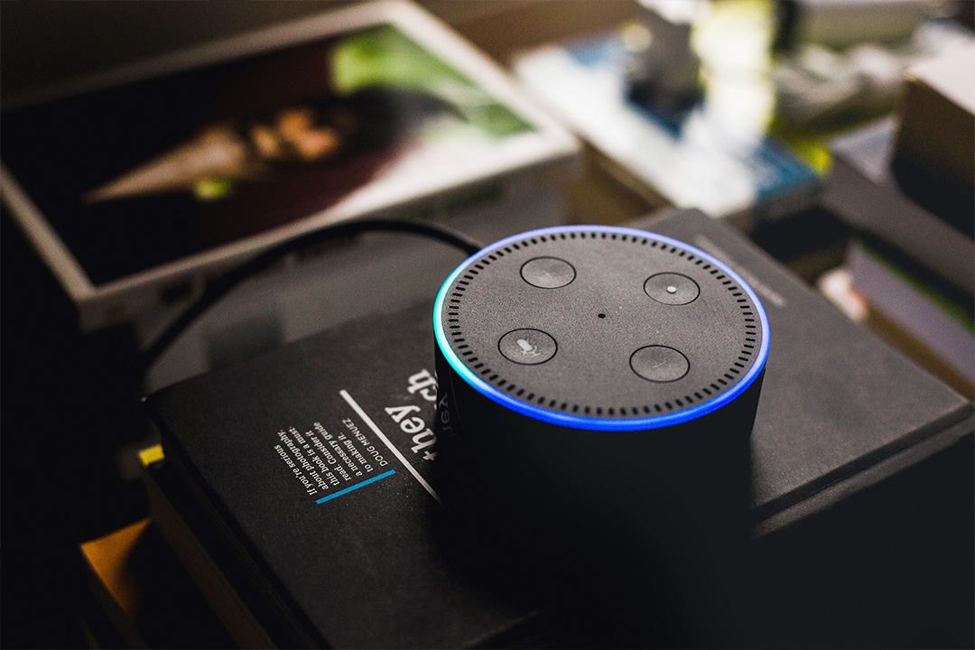 Smart speaker with a blue light ring activated, part of home security devices, resting on a dark surface surrounded by various electronics and printed photos.