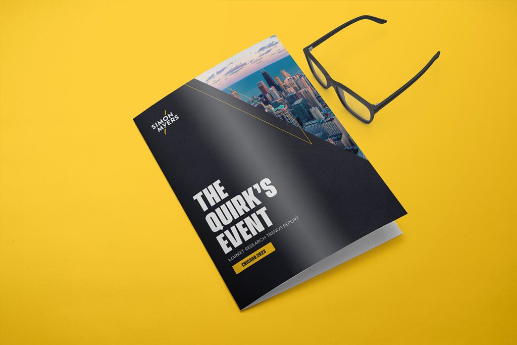 A brochure for "the quirks event" lies on a vibrant yellow surface, partially covered by a pair of black nerd glasses. The cover features a cityscape with skyscrapers.