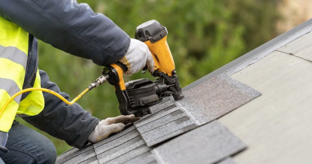 A professional installer in a reflective vest and gloves uses a nail gun on shingles while repairing a roof.