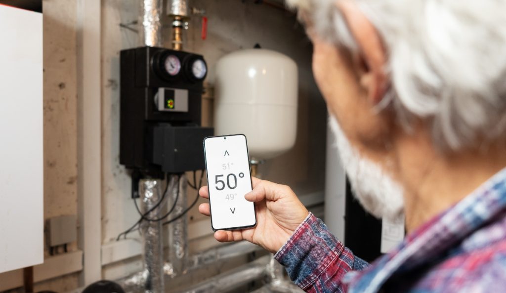 An elderly person monitors a heating system using a smartphone app that displays temperature and voltage, in a utility room, featuring recommendations from top home improvement brands.