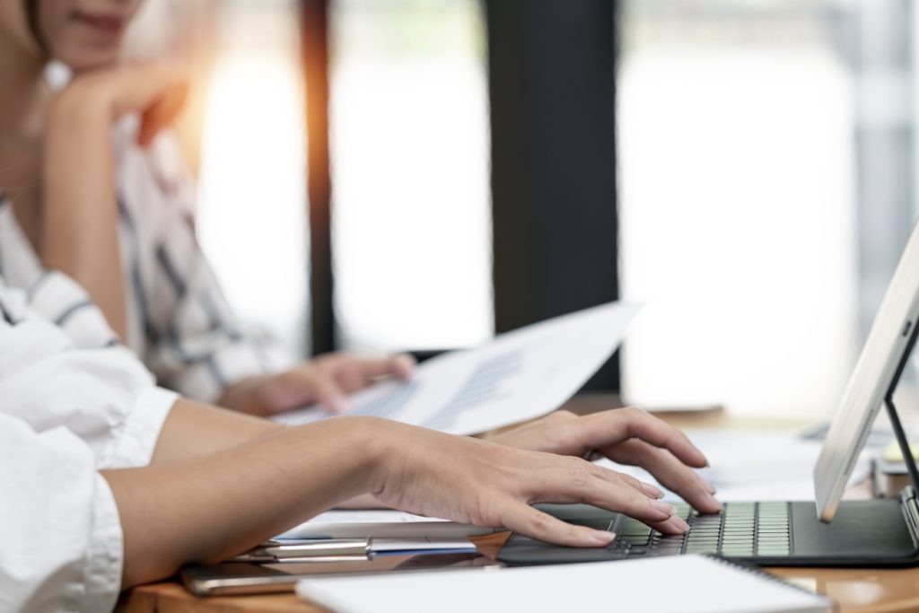 Close-up of a person's hands typing on a laptop keyboard, focusing on digital marketing strategies, with another person holding documents in the background, both sitting at a wooden desk in a well-lit office