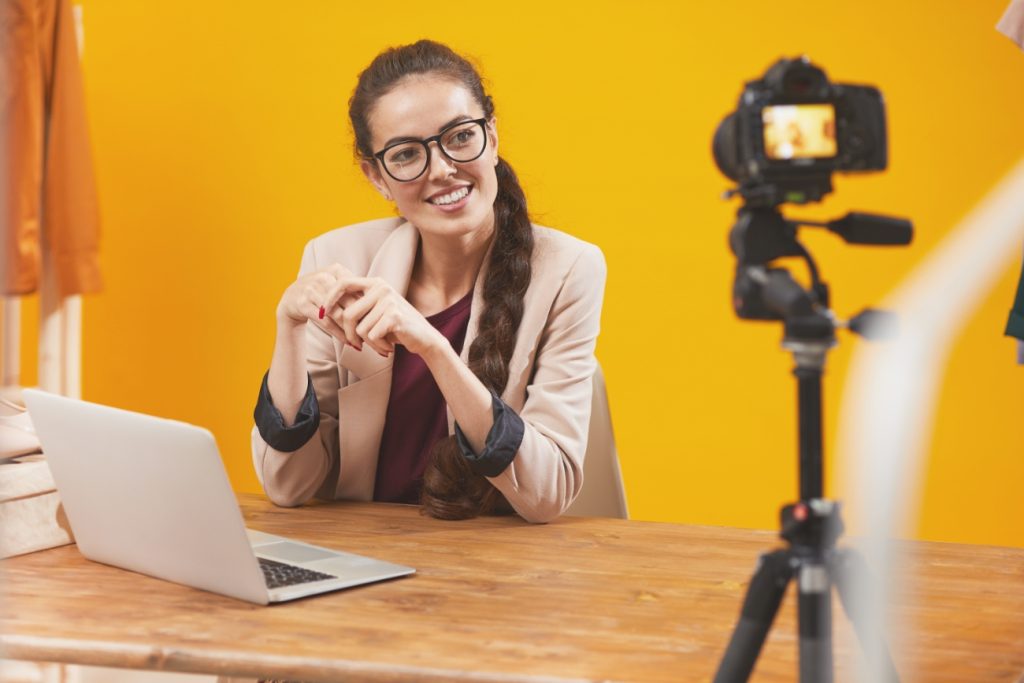 A smiling woman with glasses, wearing a blazer, sits at a desk with a laptop, recording a digital marketing video on a camera against a yellow background.