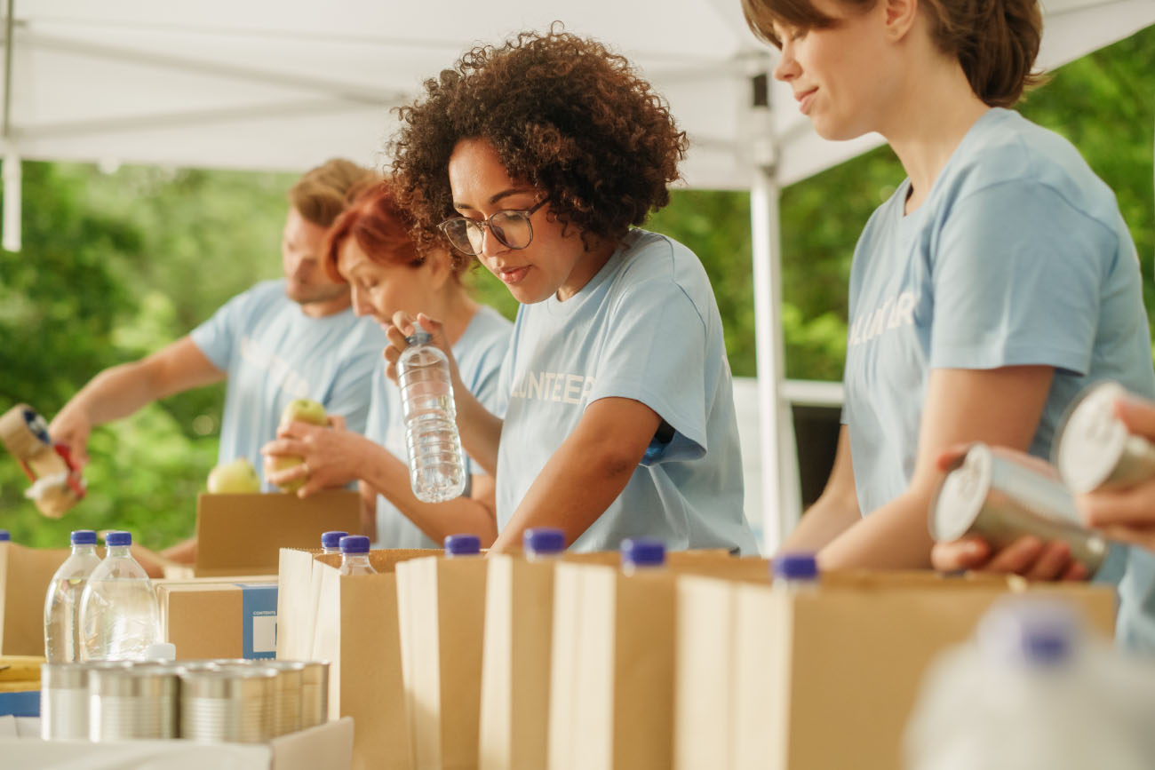 A group of volunteers in blue shirts preparing supplies at a corporate giving event, focusing on assembling packages under a white tent outdoors.