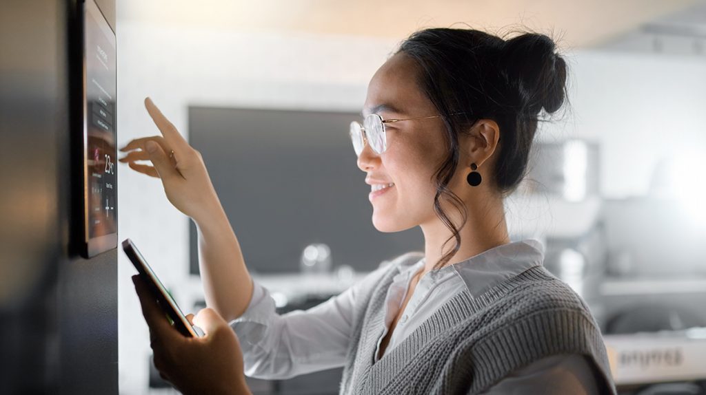 A smiling woman with glasses uses a smartphone in one hand while interacting with a digital wall panel in a modern kitchen setting, reflecting the latest trends of the homebuilding boom.