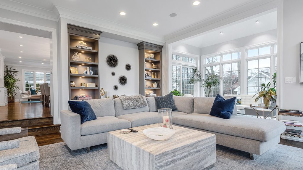 A spacious, modern living room with a large grey sectional sofa, a coffee table, built-in bookshelves, and large windows providing ample natural light, reflecting current real estate trends.