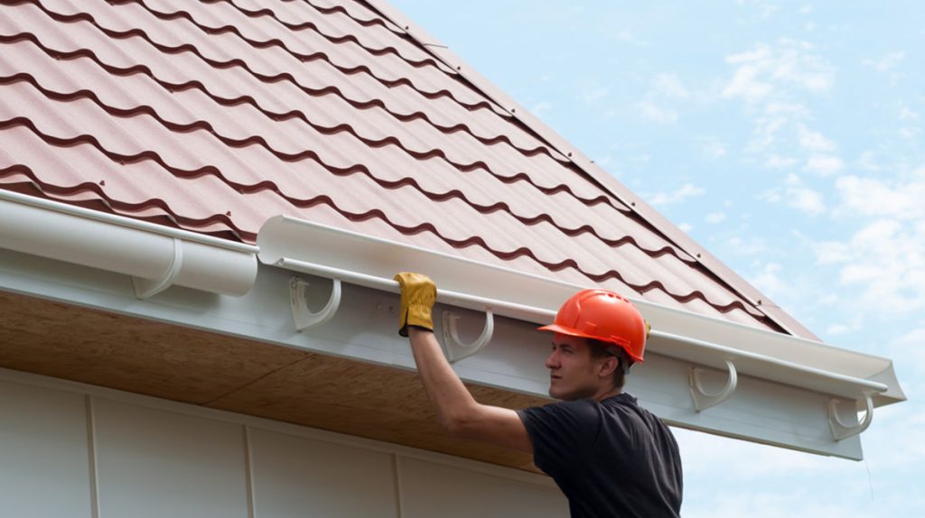 A man in a red hard hat and yellow gloves installs a white gutter on a house with a red tiled roof under a clear blue sky, demonstrating building industry trends.