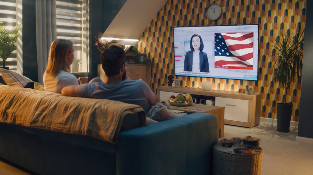 Two people sit on a sofa in a dimly lit living room, watching a news broadcast on TV. The screen shows a news reporter with an American flag in the background, discussing political advertising. The room has a cozy ambiance with patterned wallpaper, houseplants, and a clock above the TV.