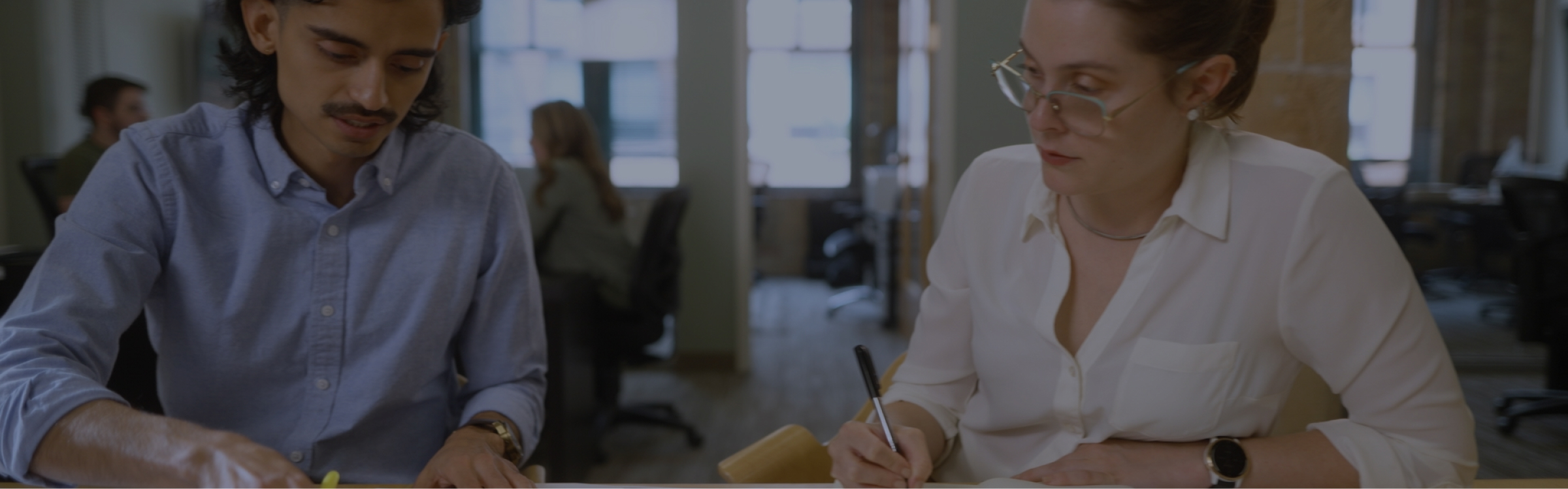 Two individuals are seated at a table in an office environment, both engaged in writing or reviewing documents. The person on the left wears a light blue shirt, and the person on the right wears a white blouse and glasses. Other office workers are visible in the background.