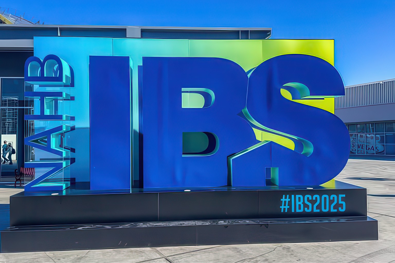 A large 3D sign featuring "IBS" in blue letters alongside "NAHB" stands prominently against a green-to-blue gradient backdrop. The hashtag "#IBS2025" graces the lower right corner, showcasing the industry's laser focus on sustainability at this vibrant outdoor event on a sunny day.