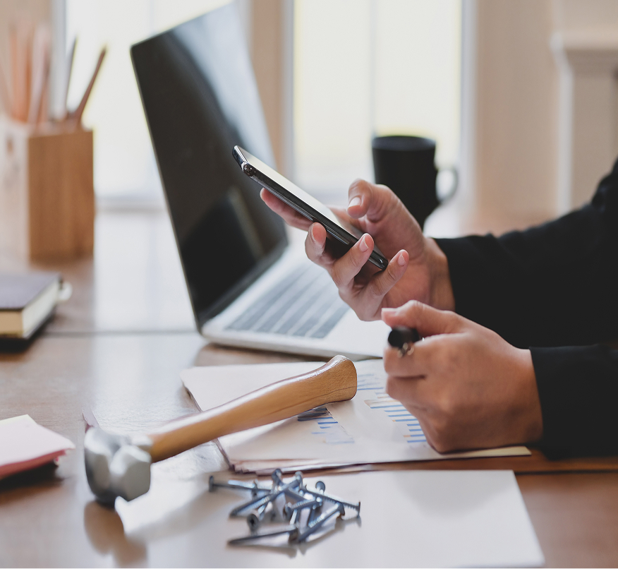 A person at a desk uses a smartphone while holding a pen. There is a laptop, documents, a hammer, and nails on the desk, blending work and DIY tools in a bright, home office setting.