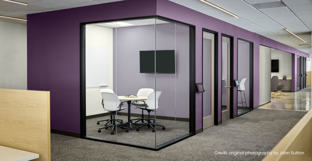 A modern office with purple walls—reflecting the latest paint trends—features a glass-walled meeting room with a small table and four white chairs. Nearby, individual office doors line the hallway. The space is bright and neatly organized.