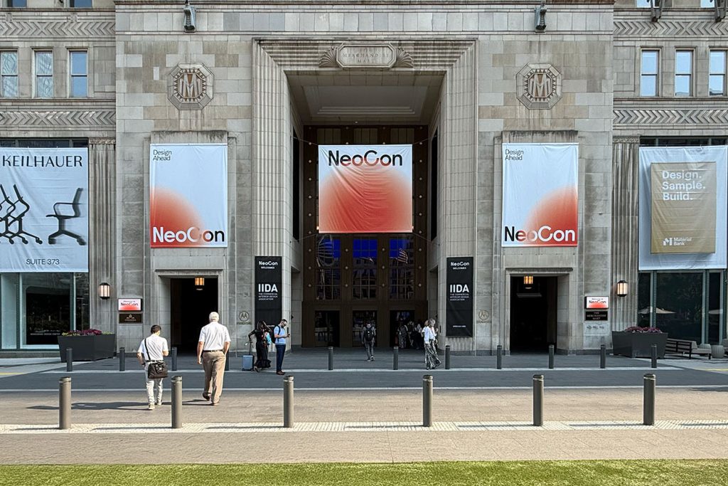 People walk toward the entrance of a grand building with banners reading "NeoCon" and "Design, Sample, Build." Under the sunny sky, attendees gather to explore special edition paint trends inside the elegant stone facade.