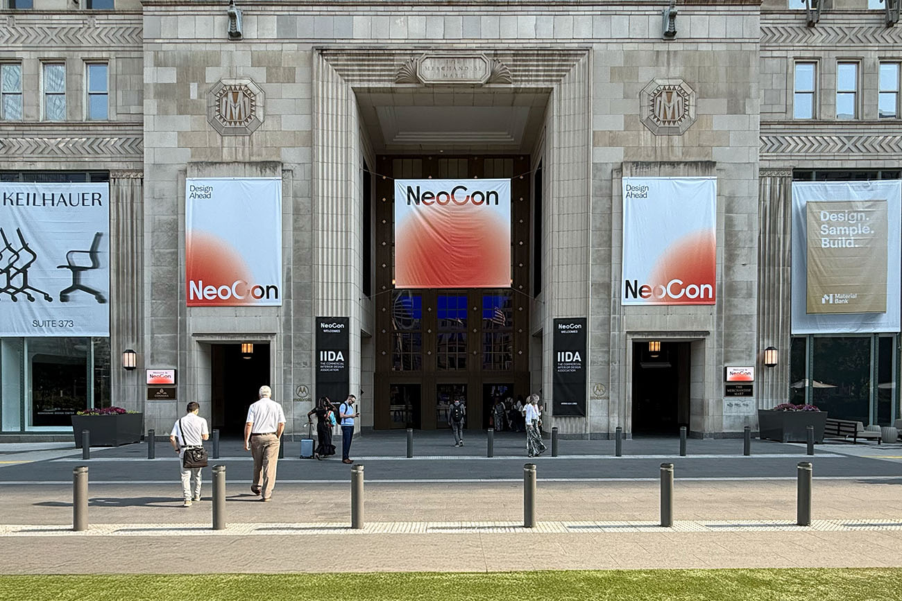 People walk toward the entrance of a grand building with banners reading "NeoCon" and "Design, Sample, Build." Under the sunny sky, attendees gather to explore special edition paint trends inside the elegant stone facade.