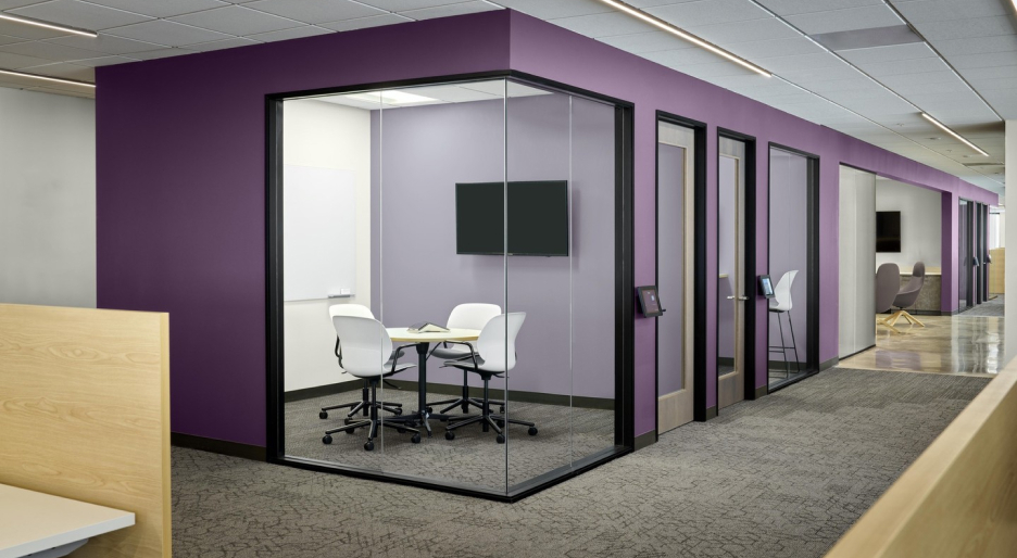 A modern office space with a purple-walled glass meeting room, inspired by the latest paint trends, contains a table and four white chairs. Adjacent are closed office doors, with open workstations and seating areas nearby.