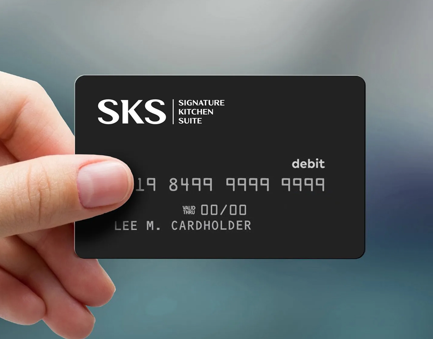 A hand holds a black debit card with the text "SKS Signature Kitchen Suite," a chip, and sample card details, including the name "LEE M. CARDHOLDER," against a blurred background.