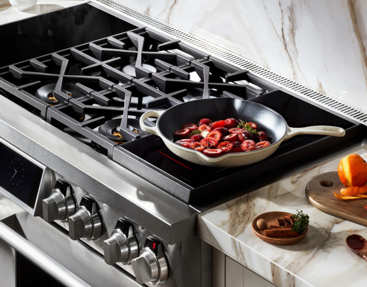 A stovetop with a cast iron skillet cooking tomatoes and herbs, next to a chopping board with orange peels and small bowls, set on a marble kitchen counter.