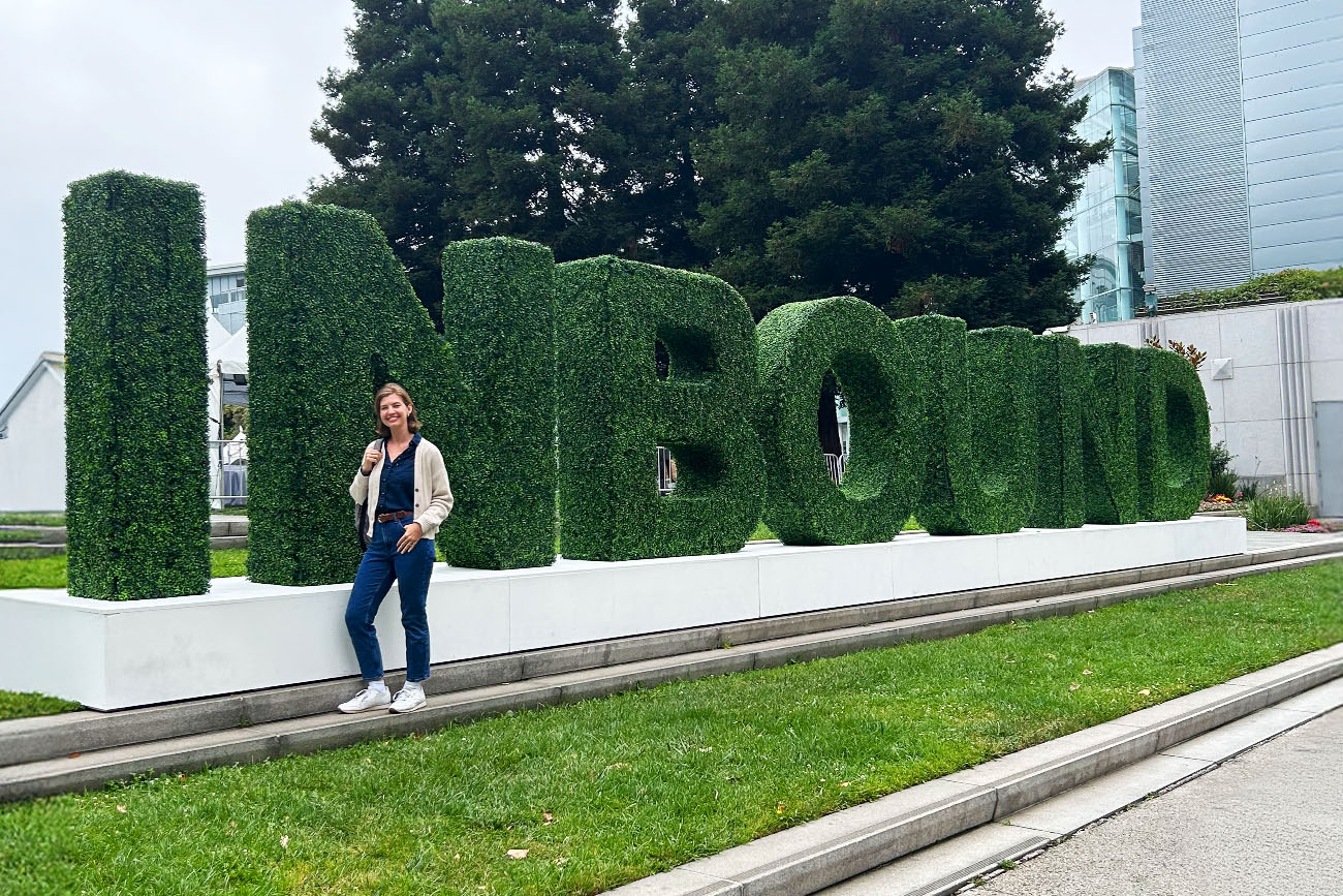 A person stands smiling next to large topiary letters spelling "INBOUND" on a sidewalk, showcasing a vibrant scene perfect for B2B marketing or home and building brands, with green grass, trees, and modern buildings in the background.