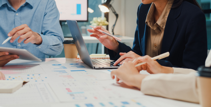 Three people sit at a desk with charts, graphs, a laptop, and tablets, discussing business data and analysis. Coffee cups and office equipment are also visible on the table.