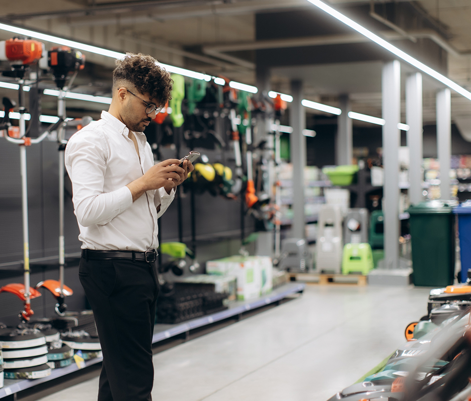 A man in a white shirt and black pants stands in a store aisle, looking at his phone. Shelves around him display various power tools and equipment. The setting appears bright and organized.