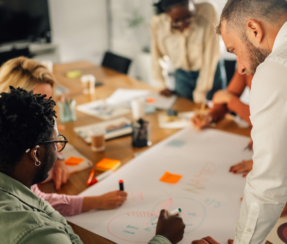Four people gather around a large table covered with paper, markers, and sticky notes, collaborating on a project. Two people are writing and drawing diagrams while others engage in discussion.
