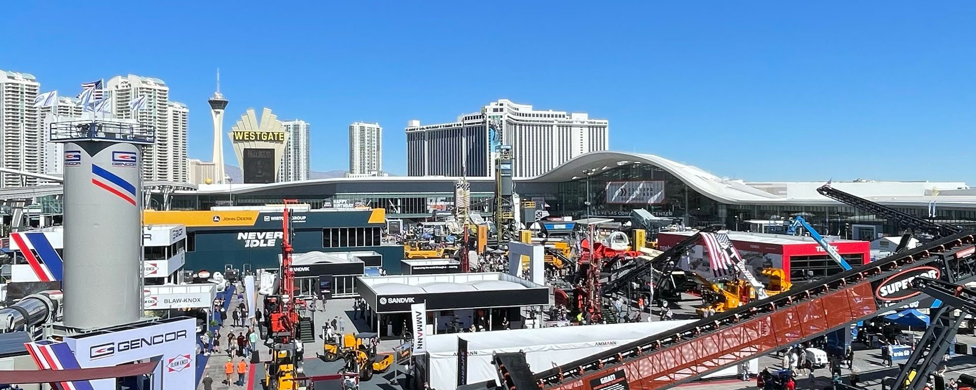 Outdoor convention with crowds, large construction equipment, and exhibitor booths under a clear blue sky. Tall hotels and the Stratosphere Tower stand in the background as ConExpo 2026 showcases innovations for smarter safety in an urban setting.