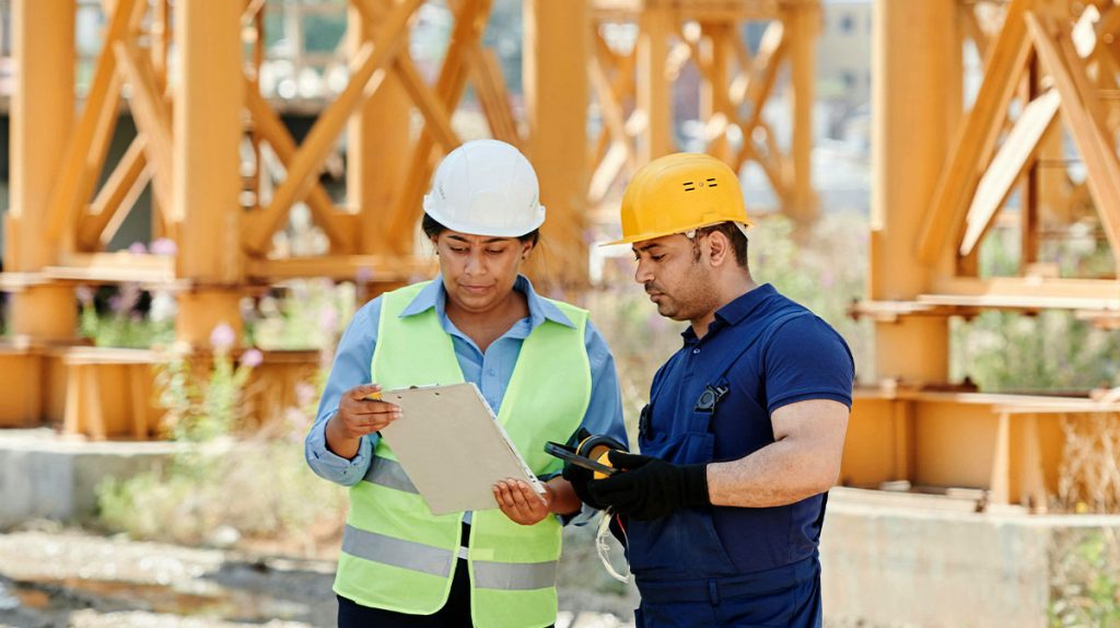 Two construction workers wearing safety gear and hard hats stand on-site. One holds a clipboard, and the other holds a tablet as they discuss Smarter Safety practices in front of a large yellow structural framework.