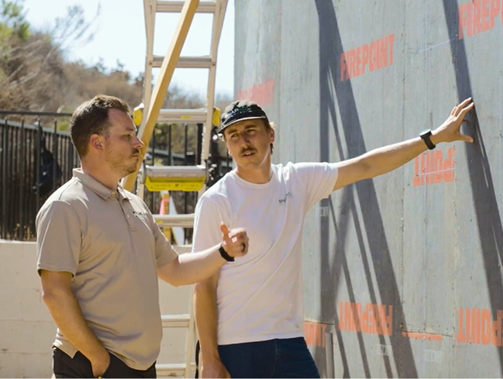 Two men stand next to a wall covered in building wrap. One man gestures while talking, and the other listens with his arm resting on the wall. A ladder and some trees are visible in the background.